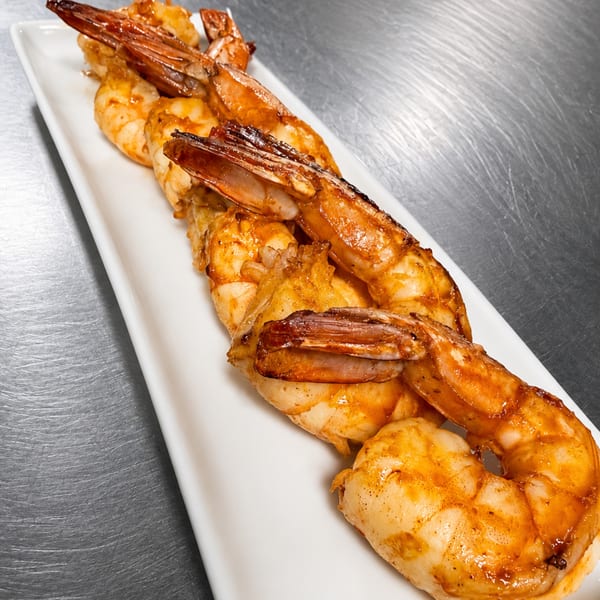 Grilled shrimp with tails on, arranged on a long white rectangular plate against a metal background.